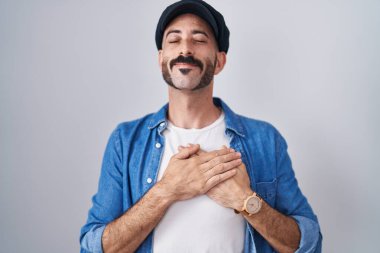 Hispanic man with beard standing over isolated background smiling with hands on chest with closed eyes and grateful gesture on face. health concept. 