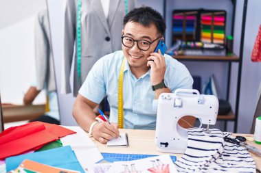 Young chinese man tailor talking on smartphone writing on notebook at tailor shop
