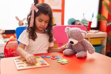 Adorable hispanic girl playing with maths puzzle game sitting on table at kindergarten