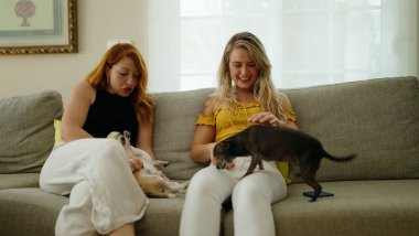 İki kadın koltukta otururken kendine güvenerek gülümsüyor. Chihuahualar evde.