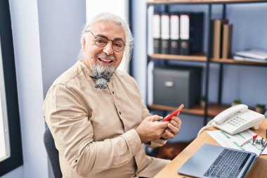 Ofiste dizüstü bilgisayar ve akıllı telefon kullanan orta yaşlı, kır saçlı bir iş adamı.