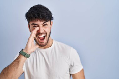 Hispanic man with beard standing over white background shouting and screaming loud to side with hand on mouth. communication concept. 