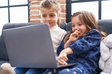 Evdeki kanepede oturan sevimli kızlar dizüstü bilgisayar kullanıyorlar.