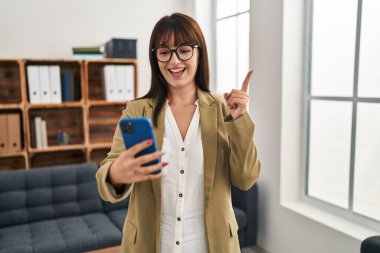 Ofiste çalışan genç esmer kadın akıllı telefonuyla gülümsüyor mutlu bir şekilde elini ve parmağını yana doğru gösteriyor. 