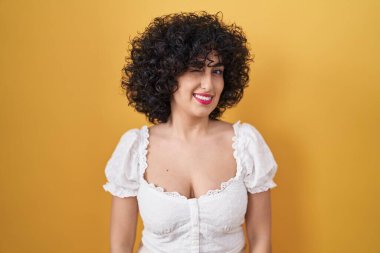 Sarı arka planda kıvırcık saçlı genç esmer kadın kameraya seksi bir ifadeyle bakıyor, neşeli ve mutlu bir yüzle.. 