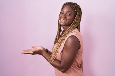 Pembe arka planda örülü saçları olan Afro-Amerikalı bir kadın avuç içi açık bir şekilde fotokopi alanını gösteriyor, gülümseyerek mutlu bir gülümseme sunuyor. 