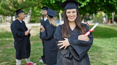 Bir grup öğrenci üniversite kampüsünde kollarını kavuşturarak diploma alarak mezun oldular.