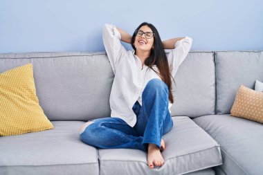 Young hispanic woman relaxed with hands on head sitting on sofa at home