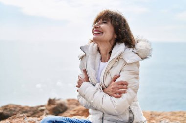 Orta yaşlı bir kadın kollarını kavuşturup deniz kenarında nefes alıyor.