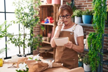 Middle age woman florist talking on smartphone reading notebook at flower shop
