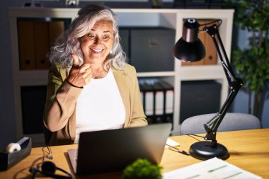 Gri saçlı orta yaşlı bir kadın gece geç saatlerde bilgisayar bilgisayarı kullanarak mutlu bir el hareketi yapıyor. Kameraya bakıp başarıyı gösteren ifade. 