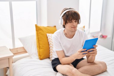 Sarışın genç adam dokunmatik yastıkta video izliyor yatak odasında oturuyor.