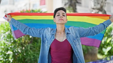 Parkta gökkuşağı bayrağı taşıyan, ciddi bir ifadeyle duran genç, güzel İspanyol kadın.