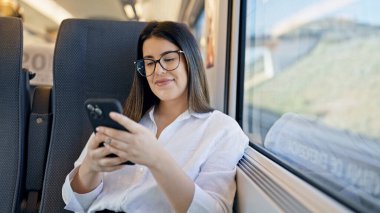 Genç, güzel, İspanyol bir kadın gülümsüyor. Akıllı telefon kullanıyor. Trende oturuyor.