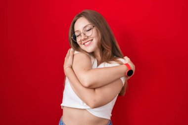 Kafkasyalı genç bir kadın kırmızı arka planda durup kendine sarılıyor mutlu ve pozitif, kendinden emin gülümsüyor. Sevgi ve özgüven. 