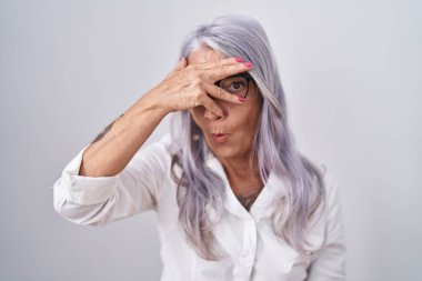 Beyaz arka planda duran gözlüklü orta yaşlı bir kadın. Şok olmuş bir şekilde yüzünü ve gözlerini kapatıyor. Utanmış bir ifadeyle parmaklarının arasından bakıyor.. 