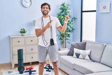 Evde spor giyinmiş yakışıklı Latin adam gülümsüyor ve kameraya bakıyor. İki elini ve parmaklarını kenarı işaret ediyor.. 