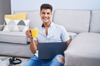 Genç İspanyol adam dizüstü bilgisayar kullanıyor ve evde oturmuş kahve içiyor.