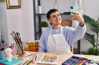 Genç ikili olmayan sanatçı gülümsüyor. Sanat stüdyosundaki akıllı telefondan özçekim yapıyor.