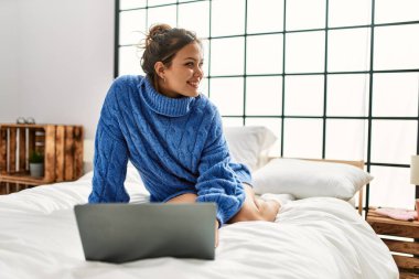 Young beautiful hispanic woman using laptop sitting on bed at bedroom
