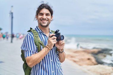 Young hispanic man tourist wearing backpack using professional camera at seaside