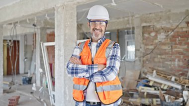 Orta yaşlı inşaatçı kendinden emin gülümsüyor inşaat alanında kollarını kavuşturmuş duruyor.