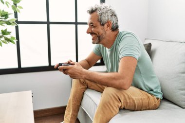 Middle age grey-haired man playing video game sitting on sofa at home