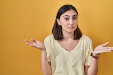Sarı arka plan üzerine sıradan tişörtler giyen İspanyol bir kız. Kolları ve elleri havada, kafası karışmış bir ifade. şüphe kavramı. 