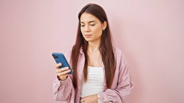 Genç, güzel İspanyol bir kadın, izole edilmiş pembe arka planda ciddi bir ifadeyle akıllı telefon kullanıyor.