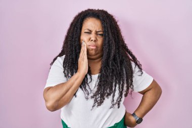 Artı, diş ağrısı ya da diş ağrısı yüzünden pembe arka planda dikilen İspanyol kadın ellerinin acı dolu ifadesiyle dudaklarına dokunuyor. dişçi 