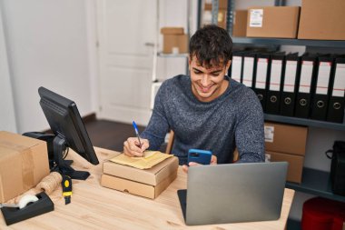 Young hispanic man ecommerce business worker using smartphone writing on package at office