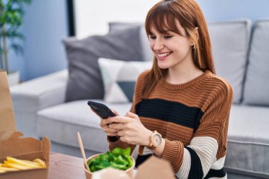 Akıllı telefon kullanan genç bir kadın evde yemek yiyor.