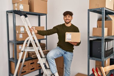 Young arab man ecommerce business worker holding package standing on ladder at office