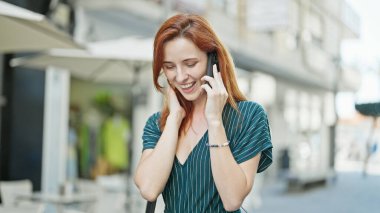 Genç kızıl saçlı, gülümseyen, kendine güvenen bir kadın kahve dükkanının terasında akıllı telefondan konuşuyor.