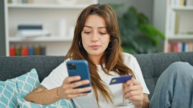 Genç, İspanyol bir kadın akıllı telefon ve kredi kartıyla alışveriş yapıyor. Evdeki koltukta oturuyor.
