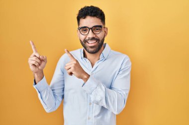 Hispanic man with beard standing over yellow background smiling and looking at the camera pointing with two hands and fingers to the side. 