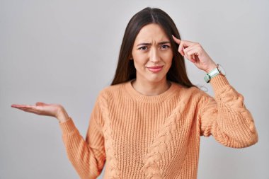 Beyaz arka planda duran esmer bir kadın avuç içinin açık olmasından rahatsız ve kafası karışık bir şekilde fotokopi alanını gösteriyor ve alnını işaret ediyor. Bunu bir düşün.. 