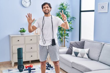 Yakışıklı Latin adam spor kıyafetlerini giyer ve yedinci parmaklarıyla gösterip işaret ederken kendinden emin ve mutlu gülümser.. 