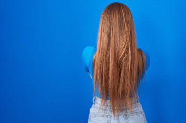 Kızıl saçlı kadın mavi arka planda dikiliyor. Arkaya bakıyor ve kollarını kavuşturuyor. 