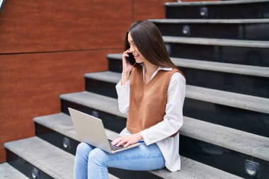Young beautiful hispanic woman talking on smartphone using laptop sitting on stairs at street