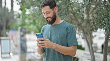 Genç İspanyol adam parkta gülümseyen akıllı telefon kullanıyor.