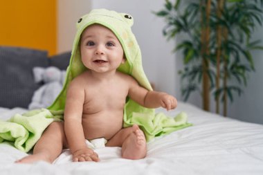 Kurbağa bornozu giymiş beyaz bebek yatak odasında yatakta oturuyor.