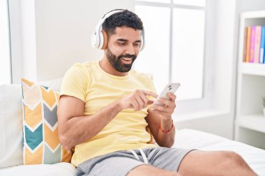 Young arab man listening to music sitting on bed at bedroom