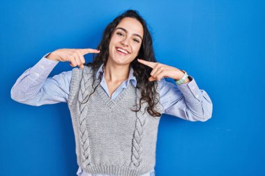 Genç esmer kadın mavi arka planda durup neşeli bir şekilde gülümseyerek parmaklarını ve ağzını işaret ediyor. diş sağlığı kavramı. 