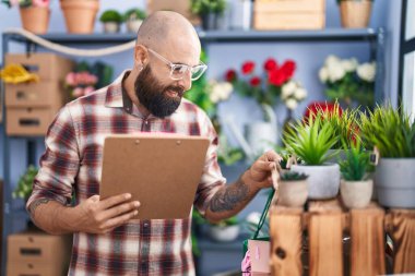 Young bald man florist reading document holding plant at flower shop