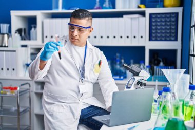 Laboratuvarda kan testi tüpü tutan dizüstü bilgisayar kullanan genç Latin bir bilim adamı.
