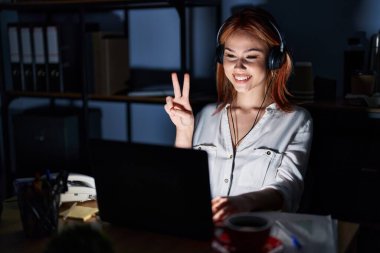 Young caucasian woman working at the office at night showing and pointing up with fingers number two while smiling confident and happy. 