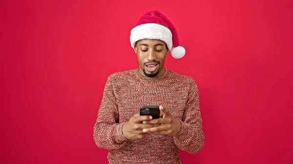 Afrikanischer Mann Mit Weihnachtsmütze Und Smartphone Lächelt Über Isolierten Roten — Stockfoto