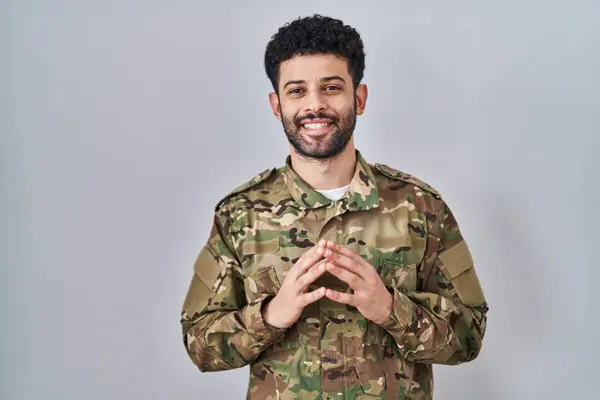 Arab man wearing camouflage army uniform hands together and fingers crossed smiling relaxed and cheerful. success and optimistic