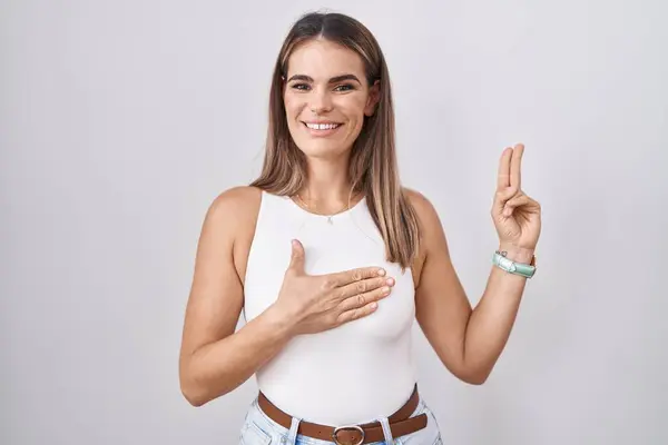 西班牙裔年轻女子站在白种人的背景上 面带微笑 双手托着胸膛和手指发誓 宣誓效忠 — 图库照片